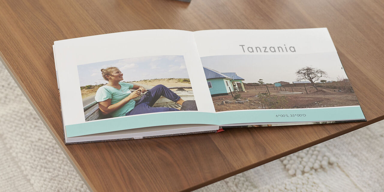 On a wooden table lies an open photo book with pictures and the headline "Tanzania". Above the photo book is a dark tray with a candle and shell. A light carpet can be seen under the table.