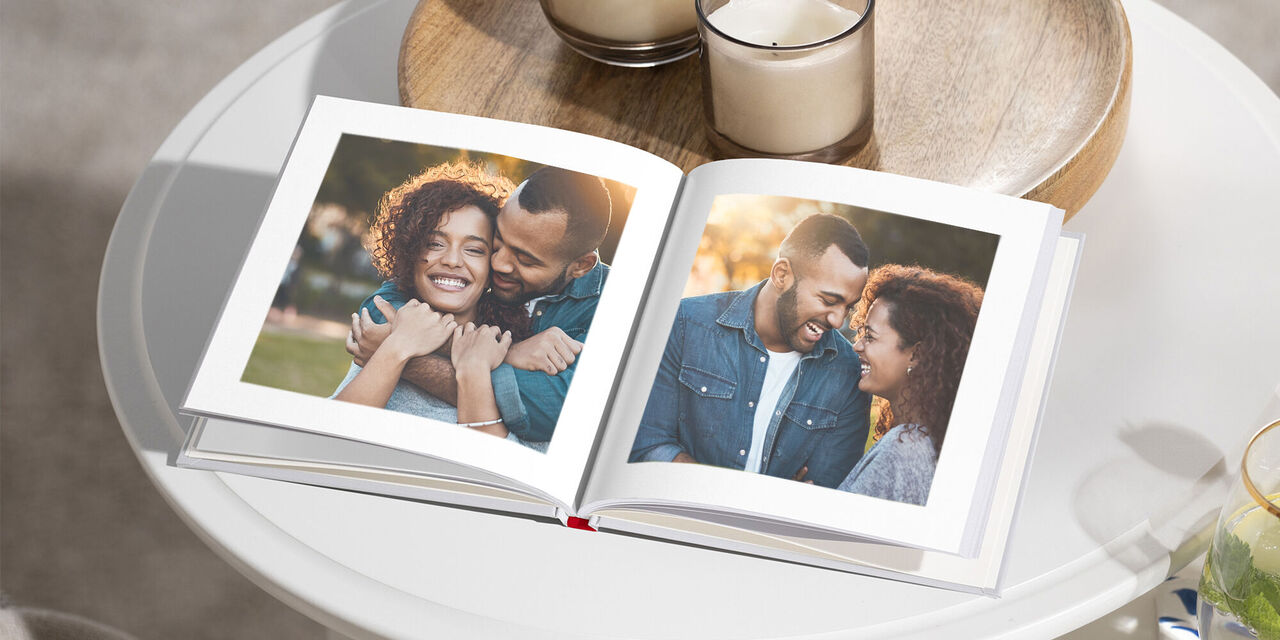 A CEWE PHOTOBOOK in Small Square format lies unfolded on a coffee table. Photos of a happy couple can be seen on the pages. On the table there is also a wooden plate with two candles and a glass of water.