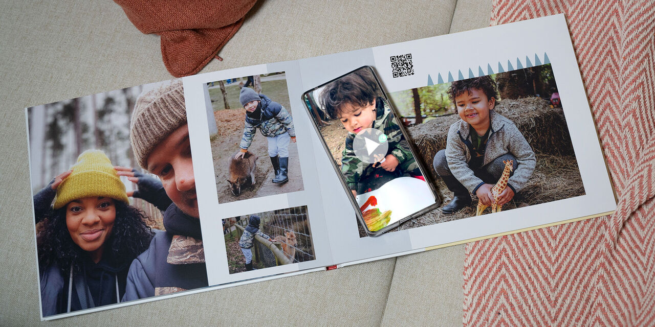 An open photo book showing family photographs printed on the pages, alongside a printed QR code. A smartphone is being used to scan the QR code in order to view an online video