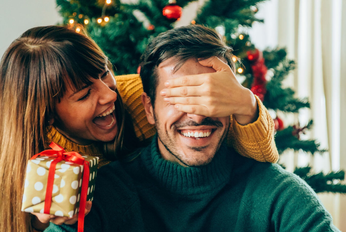 A couple embraces in front of a Christmas tree while holding a gift.