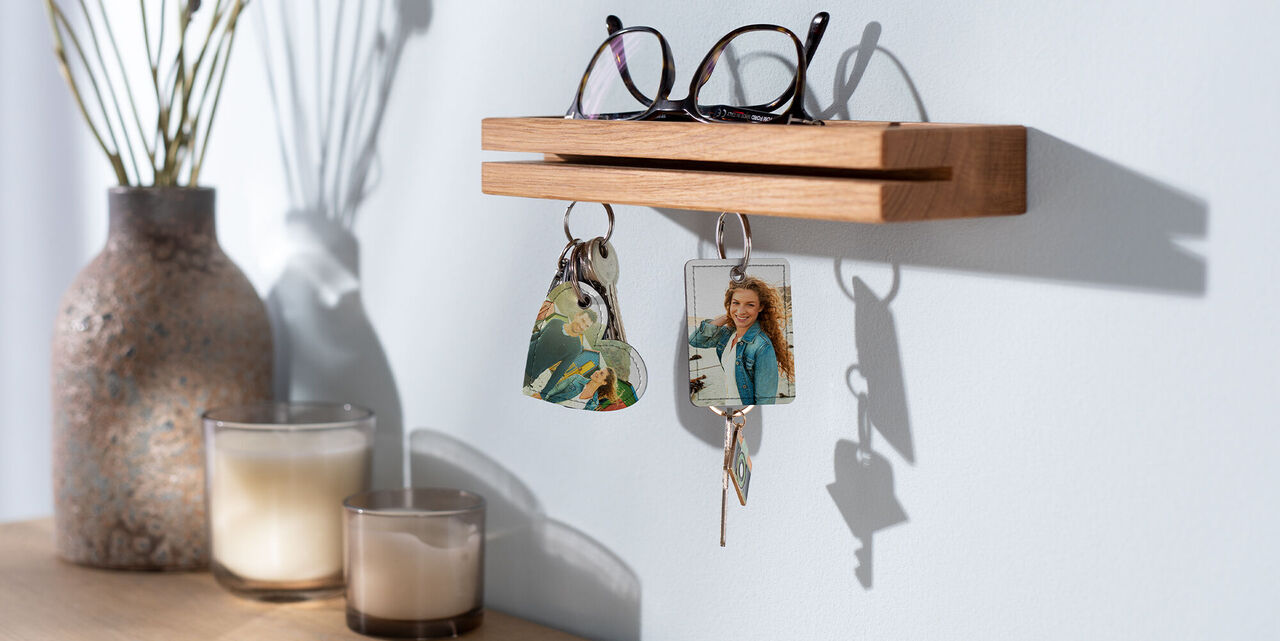 The keyrings with a photo in heart and rectangular shape hang on a key board. On the key rack lies glasses with a black frame. In the background is a vase with dried flowers and two candles.
