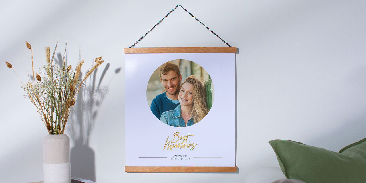A premium photo poster hangs on a Wooden Hanger on a white wall. A photo can be seen in the middle of a circle. Underneath is the slogan "Best Memories". To the left of the poster is a small table with a vase of dried flowers. On the right is a green sofa cushion.